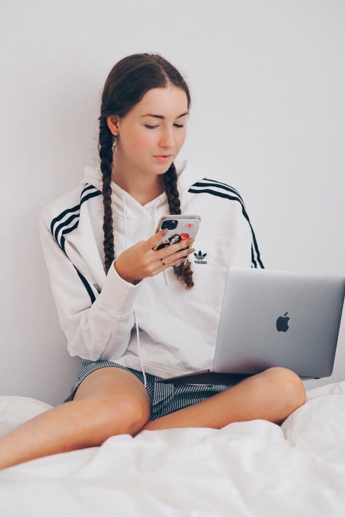 brunette woman with plaits and white adidas top and denim shorts on computer and phone