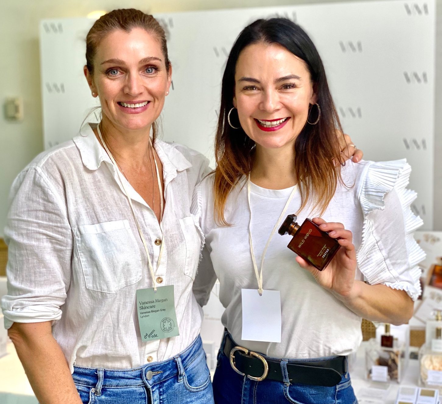 two brunette women wearing blue jeans and white shirts, one holding brown bottle of perfume