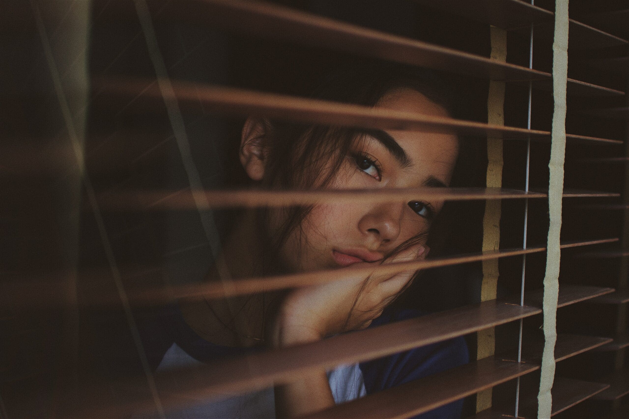 Girl looking through the blinds of her window bored