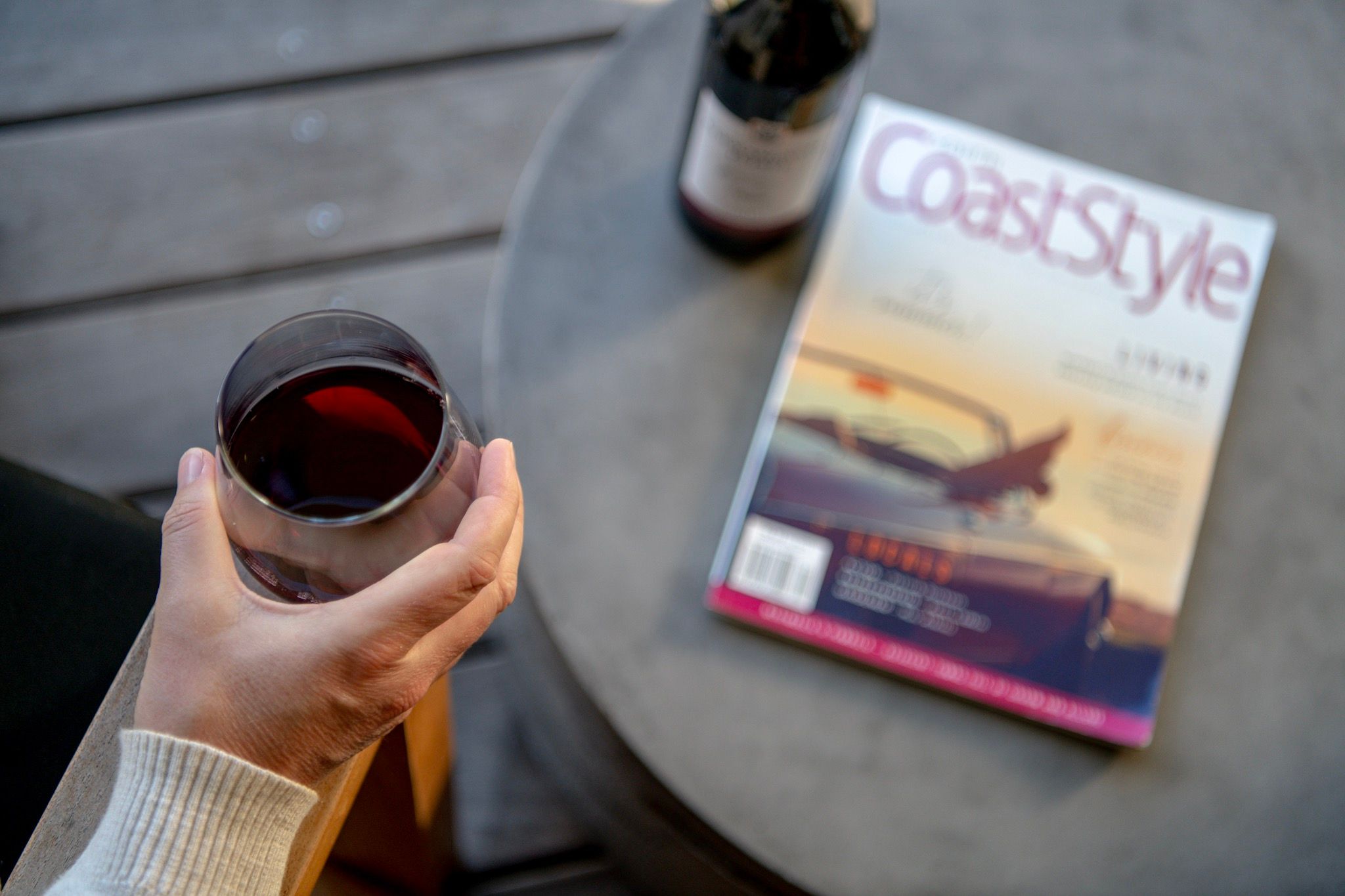 a hand holding red wine, and a magazine on table with bottle of red wine
