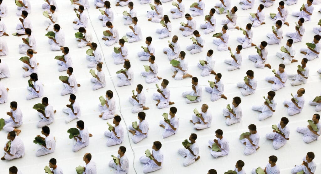 An aerial shot of male meditators in an meditation retreat dressed in white sitting on the floor and holding a green leaf. 