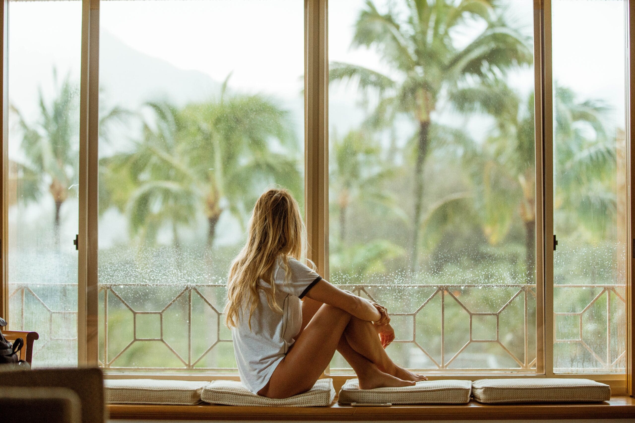 blonde woman looking out a large window at palm trees